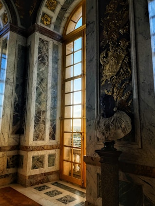 A lavish interior featuring a tall, ornate window with golden frames allowing natural light to stream in. The room is adorned with intricate, decorative marble walls and archways that include gold accents and classical motifs. A sculpted bust is placed on a pedestal, adding an element of art and history to the elegant setting.