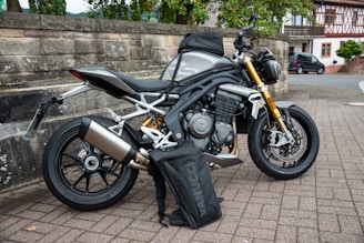 A sleek, modern motorcycle with a metallic silver body and gold front forks is parked on a paved area near a stone wall. A black bag branded 'VANUCCI' rests against the motorcycle. The background features trees, a traditional building, and a parked car, suggesting a suburban or rural setting.