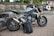 A sleek, modern motorcycle with a metallic silver body and gold front forks is parked on a paved area near a stone wall. A black bag branded 'VANUCCI' rests against the motorcycle. The background features trees, a traditional building, and a parked car, suggesting a suburban or rural setting.