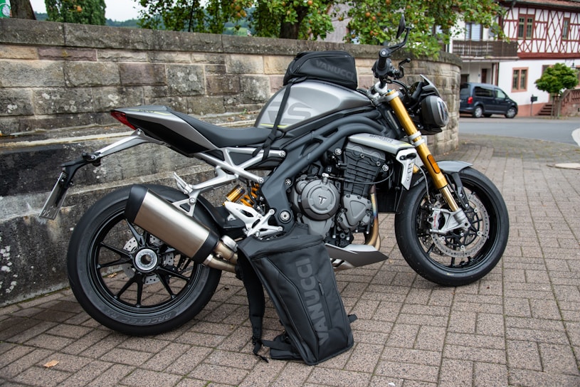 A sleek, modern motorcycle with a metallic silver body and gold front forks is parked on a paved area near a stone wall. A black bag branded 'VANUCCI' rests against the motorcycle. The background features trees, a traditional building, and a parked car, suggesting a suburban or rural setting.
