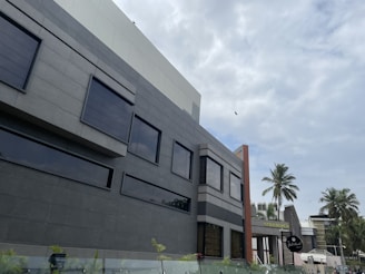 A modern building with large dark windows and a sleek facade under a partly cloudy sky. Palm trees are visible in the background alongside a few birds flying. A sign that reads 'Gold Beach' is present in the scene.