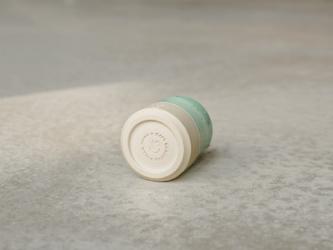 A cylindrical, light wooden cup with a smooth surface and subtle engraved text around the bottom edge lies on a textured gray concrete surface. The cup has a turquoise section with small dotted patterns near the top.