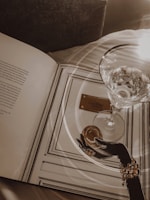 Elegant setting featuring an open book with illustrations, a hand wearing a bracelet holding a round object, and a glass filled with sparkling liquid. Soft lighting creates reflections on the glass and book, enhancing the luxurious feel.