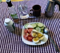 A dining setup on a purple and white checkered tablecloth includes a white plate with sliced cucumbers, tomatoes, and hard-boiled eggs seasoned with spices. There is a bottle of vinegar, a glass of water, a brown mug, a metal coffee pot, and a bottle of dietary supplements nearby.
