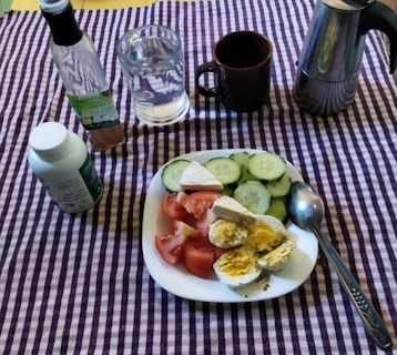 A dining setup on a purple and white checkered tablecloth includes a white plate with sliced cucumbers, tomatoes, and hard-boiled eggs seasoned with spices. There is a bottle of vinegar, a glass of water, a brown mug, a metal coffee pot, and a bottle of dietary supplements nearby.
