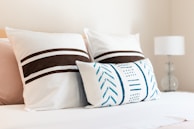 A neatly arranged bed with three decorative pillows. The two larger pillows feature brown horizontal stripes, while the smaller, rectangular pillow has blue geometric patterns. A white lamp with a clear base is visible on a bedside table to the right.