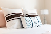A neatly arranged bed with three decorative pillows. The two larger pillows feature brown horizontal stripes, while the smaller, rectangular pillow has blue geometric patterns. A white lamp with a clear base is visible on a bedside table to the right.