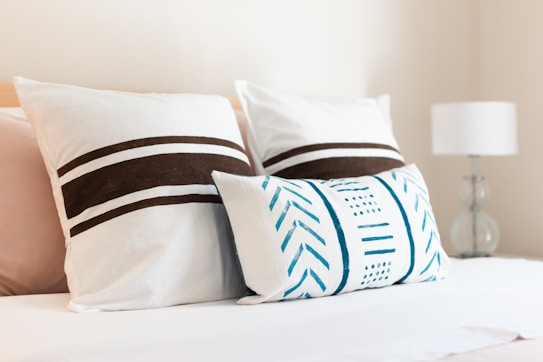 A neatly arranged bed with three decorative pillows. The two larger pillows feature brown horizontal stripes, while the smaller, rectangular pillow has blue geometric patterns. A white lamp with a clear base is visible on a bedside table to the right.