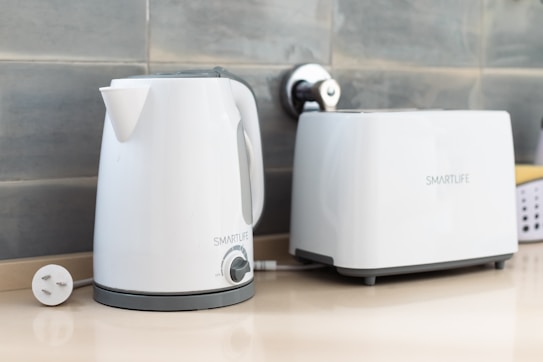 A white electric kettle and toaster are placed on a beige countertop against a tiled backsplash. Both appliances are labeled 'SMARTLIFE' and have a sleek, modern design. The kettle is plugged in, with the power cord visible.