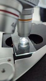 A close-up view of a microscope with a focus on the objective lens positioned above a glass slide. The microscope's metallic parts are visible, and there is a bright light illuminating the slide, highlighting its transparent surface.