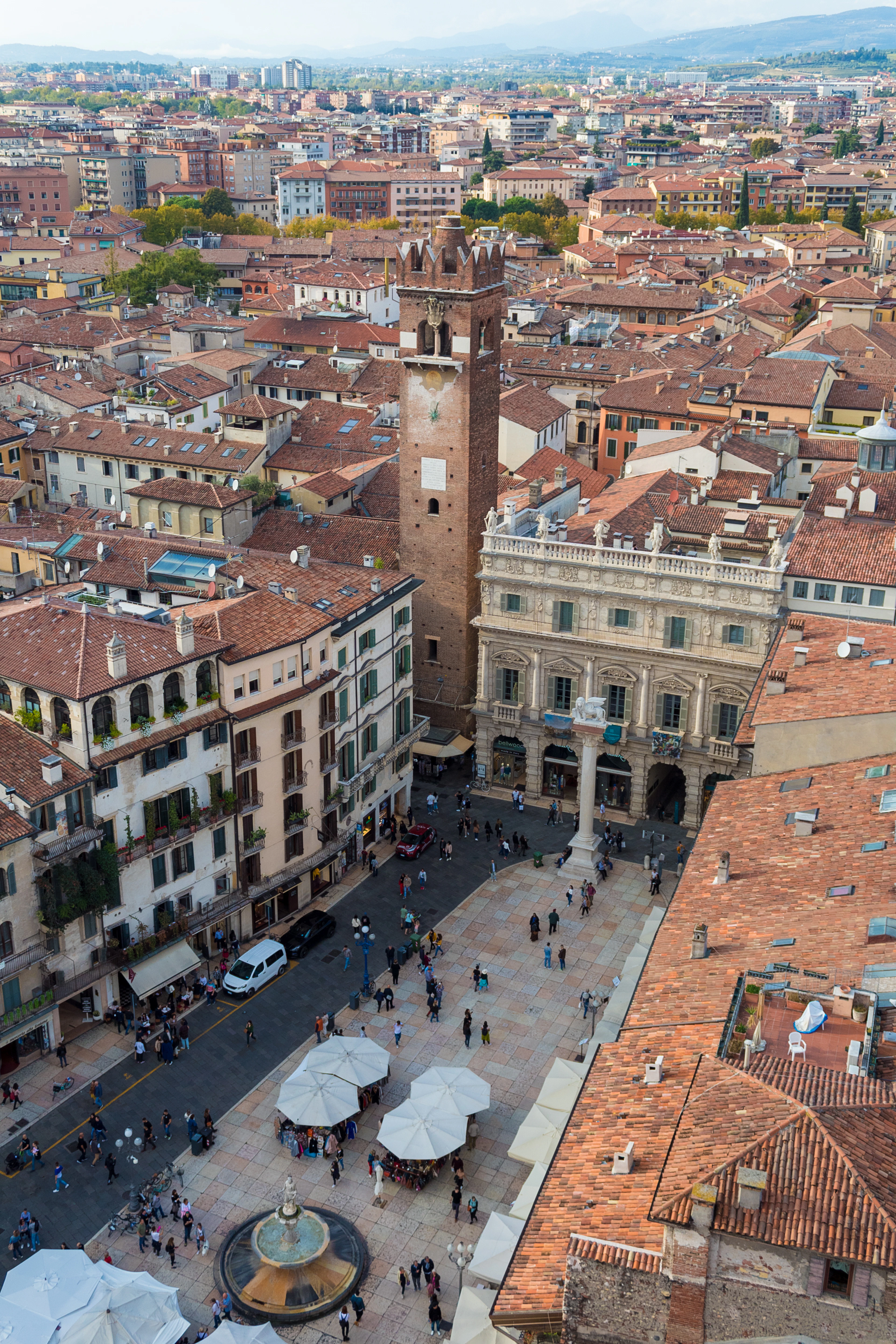 Aerial view over Piazza delle Erbe's market umbrellas and stone fountain, rooftops and a brick tower beyond.