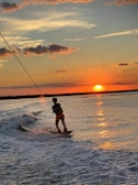 A person is wakeboarding on a body of water during sunset. The sky is painted with vibrant hues of orange and red as the sun sets on the horizon. Clouds are scattered across the sky, and the water reflects the warm colors of the sky. Waves created by a boat can be seen as the person skillfully maneuvers on the board.