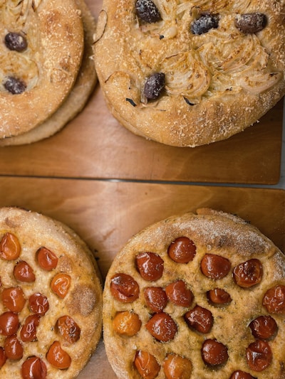 Four round focaccia breads are arranged on a wooden surface. Two are topped with caramelized onions and black olives, while the other two are covered with roasted cherry tomatoes. The bread has a rustic texture with a golden-brown crust.