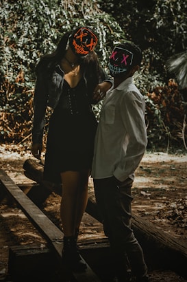 Two individuals wearing dark clothing and light-up masks stand on wooden beams in a forested area. The masks feature glowing patterns, one with red Xs and a stitched smile, and the other with pink Xs. The scene is atmospheric with dappled sunlight filtering through the trees, creating a moody environment.