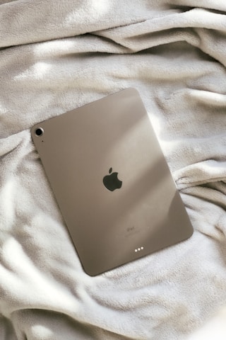 A tablet with a sleek metallic finish is resting on a fluffy, cozy blanket. The device features the Apple logo on its back, indicating it is an iPad.