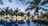 A scenic view of a tropical paradise featuring a series of palm trees surrounding an infinity pool. The horizon shows a beautiful sunset over a calm ocean. There are thatched umbrellas and lounge chairs, creating a relaxing atmosphere.