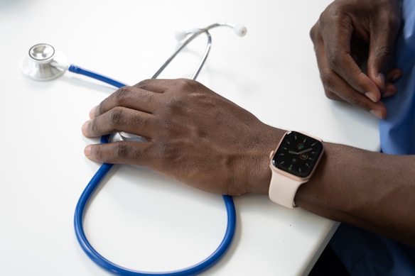 A person's hand rests on a table next to a stethoscope. The person is wearing a smartwatch on their wrist and is dressed in scrubs, suggesting a medical professional setting.