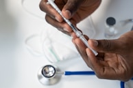 Hands holding a syringe filled with liquid, with a stethoscope and a face mask in the background on a white surface. The scene appears to be in a medical or clinical setting.