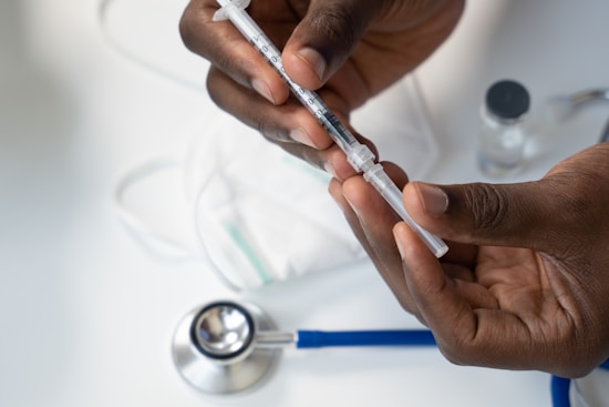 Hands holding a syringe filled with liquid, with a stethoscope and a face mask in the background on a white surface. The scene appears to be in a medical or clinical setting.