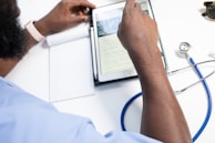 A person in a blue shirt interacts with a tablet while sitting at a desk. Nearby, a stethoscope and a pair of sunglasses are placed, suggesting a medical or clinical setting. A notepad is also partially visible, indicating note-taking or information recording.