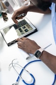 A person wearing a smartwatch writes on a notepad while using a tablet that displays medical images. A stethoscope is positioned close by on a white desk, suggesting a medical setting.
