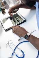 A person wearing a smartwatch writes on a notepad while using a tablet that displays medical images. A stethoscope is positioned close by on a white desk, suggesting a medical setting.