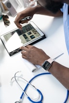 A person wearing a smartwatch writes on a notepad while using a tablet that displays medical images. A stethoscope is positioned close by on a white desk, suggesting a medical setting.