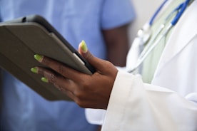 A person wearing a white coat with a stethoscope around their neck is holding a tablet device. The person's nails are painted a bright green color. Another individual in blue scrubs appears in the background, slightly out of focus.