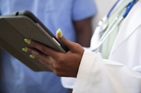 A person wearing a white coat with a stethoscope around their neck is holding a tablet device. The person's nails are painted a bright green color. Another individual in blue scrubs appears in the background, slightly out of focus.