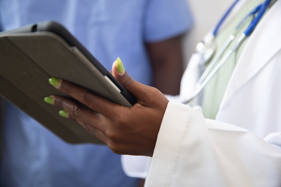 A person wearing a white coat with a stethoscope around their neck is holding a tablet device. The person's nails are painted a bright green color. Another individual in blue scrubs appears in the background, slightly out of focus.