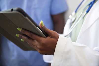 A person wearing a white coat with a stethoscope around their neck is holding a tablet device. The person's nails are painted a bright green color. Another individual in blue scrubs appears in the background, slightly out of focus.