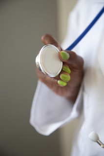 A person wearing a white coat is holding a stethoscope in their hand. The person's nails are painted a bright green color. The focus is on the stethoscope, while the background is blurred.