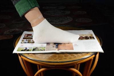 A foot wearing a white sock is placed on an open magazine resting on a round, wicker-style table. The magazine displays several colorful images and text. The person is wearing green pants, and one small skin-tone bandage can be seen on the ankle.
