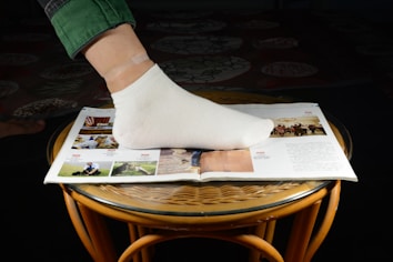 A foot wearing a white sock is placed on an open magazine resting on a round, wicker-style table. The magazine displays several colorful images and text. The person is wearing green pants, and one small skin-tone bandage can be seen on the ankle.