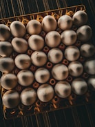 A top-down view of a tray filled with white eggs, arranged in a 6x5 grid, positioned on a textured dark surface. The eggs are placed in an orange plastic holder with circular cutouts.