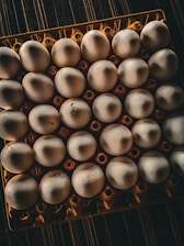 A top-down view of a tray filled with white eggs, arranged in a 6x5 grid, positioned on a textured dark surface. The eggs are placed in an orange plastic holder with circular cutouts.