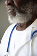 A close-up of a person with a white beard wearing a white coat and a stethoscope around their neck. The focus is on the person's lower face and upper chest.
