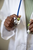 A hand with green-painted nails holding a stethoscope, which is resting against a white medical coat. The stethoscope's blue tubing is visible, and the person appears to be a healthcare professional.
