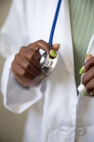 A hand with green-painted nails holding a stethoscope, which is resting against a white medical coat. The stethoscope's blue tubing is visible, and the person appears to be a healthcare professional.