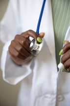 A hand with green-painted nails holding a stethoscope, which is resting against a white medical coat. The stethoscope's blue tubing is visible, and the person appears to be a healthcare professional.