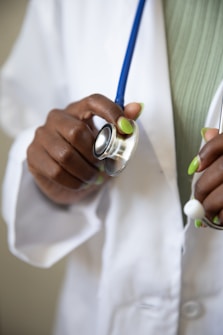 A hand with green-painted nails holding a stethoscope, which is resting against a white medical coat. The stethoscope's blue tubing is visible, and the person appears to be a healthcare professional.