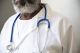A person wearing a white lab coat and a blue stethoscope around their neck. The individual has a graying beard and is wearing a white shirt underneath the lab coat.