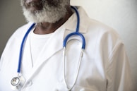 A person wearing a white lab coat and a blue stethoscope around their neck. The individual has a graying beard and is wearing a white shirt underneath the lab coat.