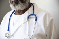 A person wearing a white lab coat and a blue stethoscope around their neck. The individual has a graying beard and is wearing a white shirt underneath the lab coat.
