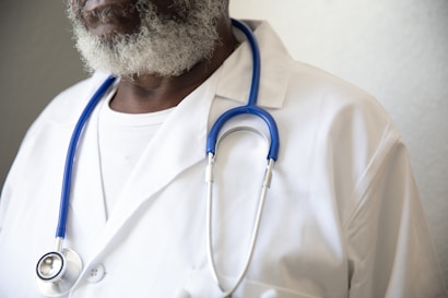 A person wearing a white lab coat and a blue stethoscope around their neck. The individual has a graying beard and is wearing a white shirt underneath the lab coat.