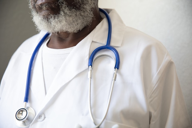 A person wearing a white lab coat and a blue stethoscope around their neck. The individual has a graying beard and is wearing a white shirt underneath the lab coat.