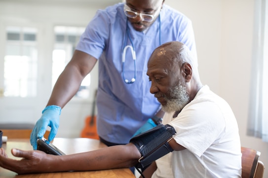 A healthcare professional wearing a blue shirt and gloves is taking the blood pressure of an elderly man who is sitting at a table. The elderly man has a thoughtful expression and is wearing a white shirt. Medical equipment is visible on the man's arm, and a stethoscope is around the healthcare professional's neck.