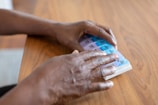 Hands are handling a weekly medication organizer placed on a wooden table. The organizer has compartments labeled with days of the week, and the colors of the compartments vary from purple to blue.