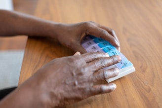 Hands are handling a weekly medication organizer placed on a wooden table. The organizer has compartments labeled with days of the week, and the colors of the compartments vary from purple to blue.