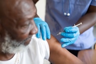 A healthcare worker wearing blue gloves is administering a vaccine to an elderly man. The scene focuses on the arm of the person receiving the injection and the hands of the healthcare worker handling the syringe.